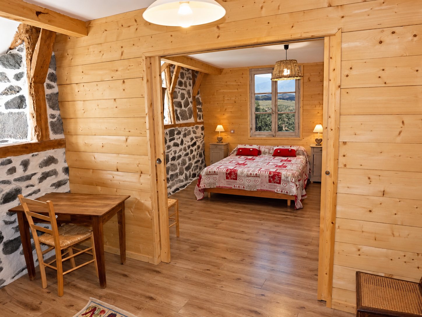 Chambre rustique aux murs en bois clair et en pierre, avec un lit double à couette rouge et blanche, deux lampes de chevet et une petite table avec chaise au premier plan.