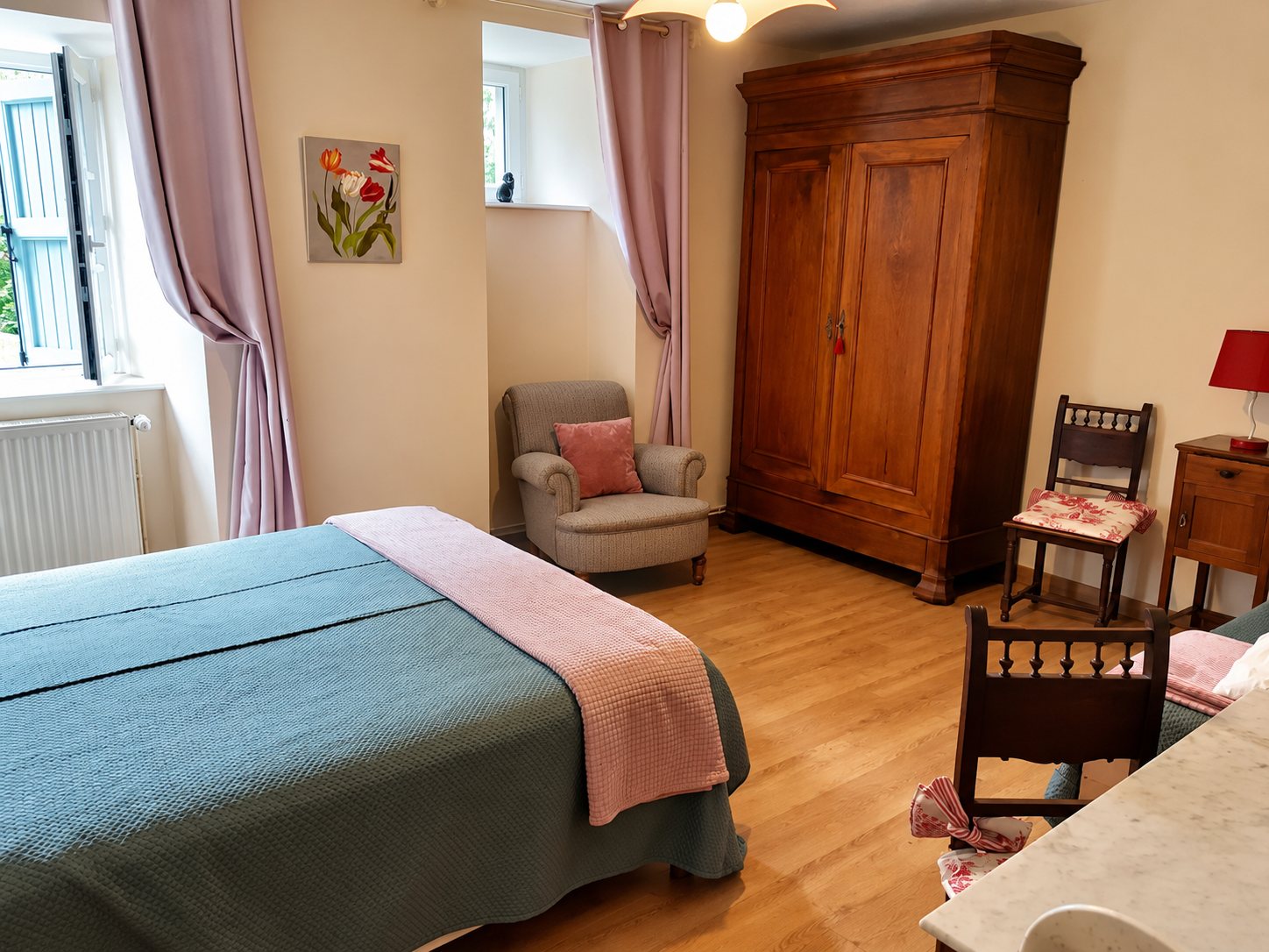 Chambre lumineuse au parquet en bois avec un lit couvert d'un plaid gris et rose, une grande armoire ancienne, un fauteuil et des rideaux mauves.