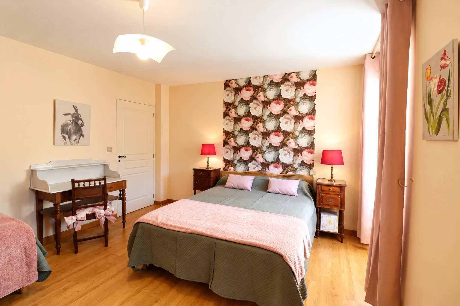 Chambre aux tons rosés avec un grand lit double, un papier peint à grosses fleurs, deux lampes rouges sur des tables de chevet en bois et un parquet clair.