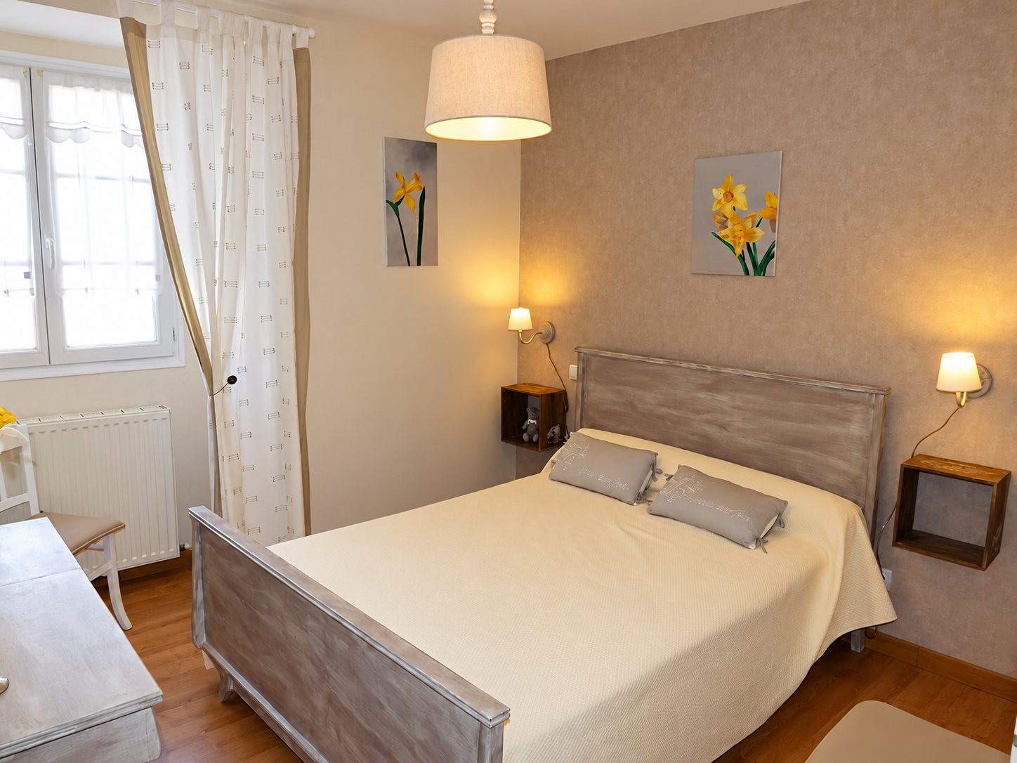 Chambre aux tons beiges avec un lit double en bois patiné, deux coussins gris, des appliques allumées et deux tableaux de jonquilles au mur.