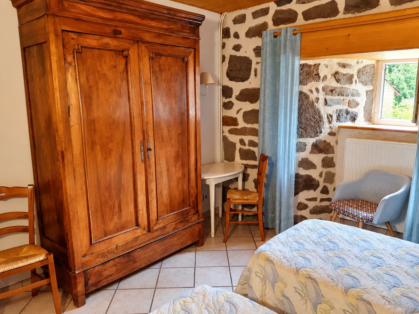 Chambre d'hôtes aux murs de pierre apparente avec une grande armoire ancienne en bois, des chaises paillées et deux lits recouverts de couvre-lits matelassés.