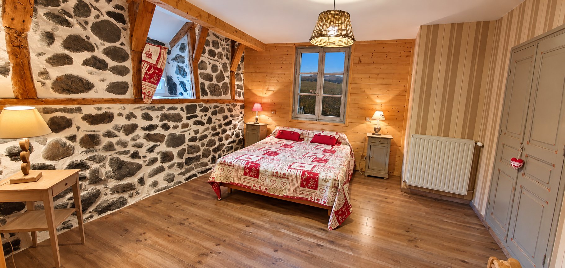 Chambre d'hôtes chaleureuse avec mur en pierres apparentes, lambris de bois clair, grand lit couvert d'un patchwork rouge et blanc et deux tables de chevet.