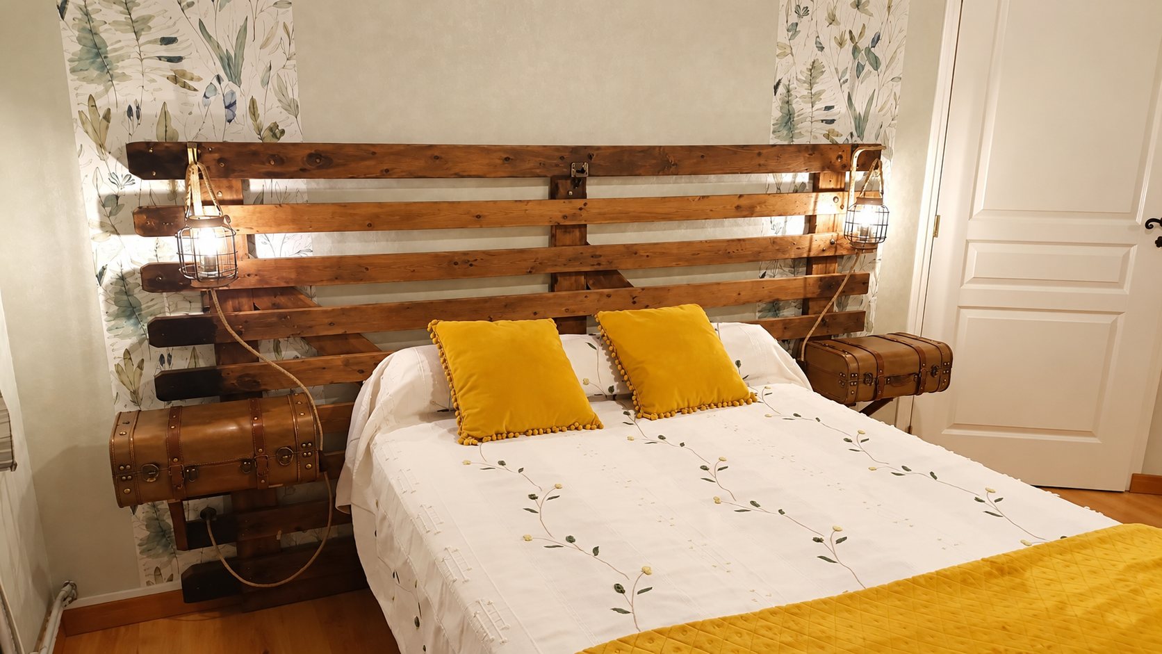 Chambre décorée d'une tête de lit en bois de palettes, de lampes cages, de valises servant de chevets et de coussins jaune moutarde sur un lit blanc.