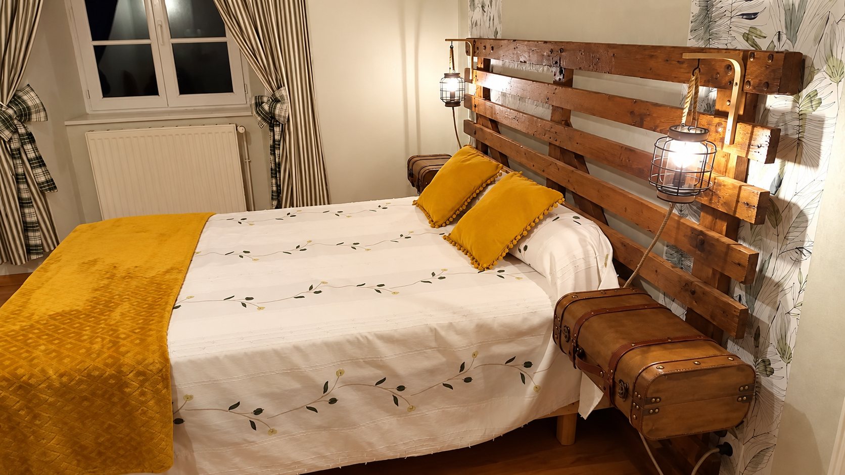 Chambre chaleureuse avec un lit orné de coussins jaune moutarde, une tête de lit en bois de palettes, des lampes cage et des valises anciennes en guise de tables de chevet.