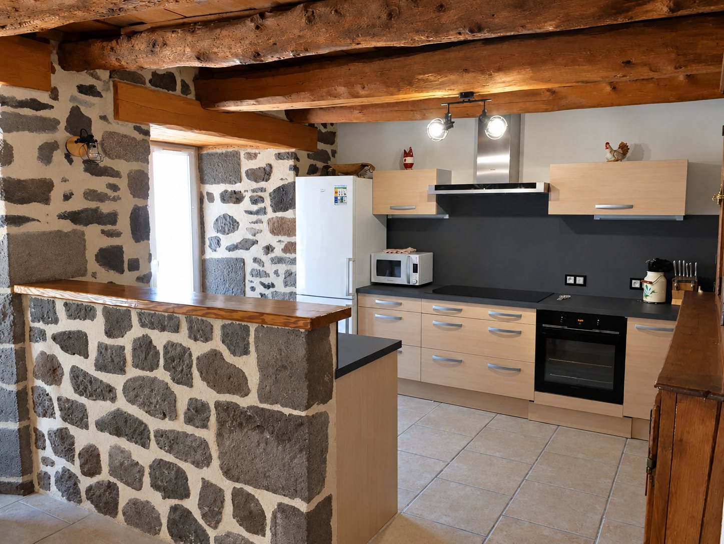 Cuisine moderne aux meubles en bois clair et plan de travail sombre, dans une pièce aux murs en pierre et aux poutres apparentes.