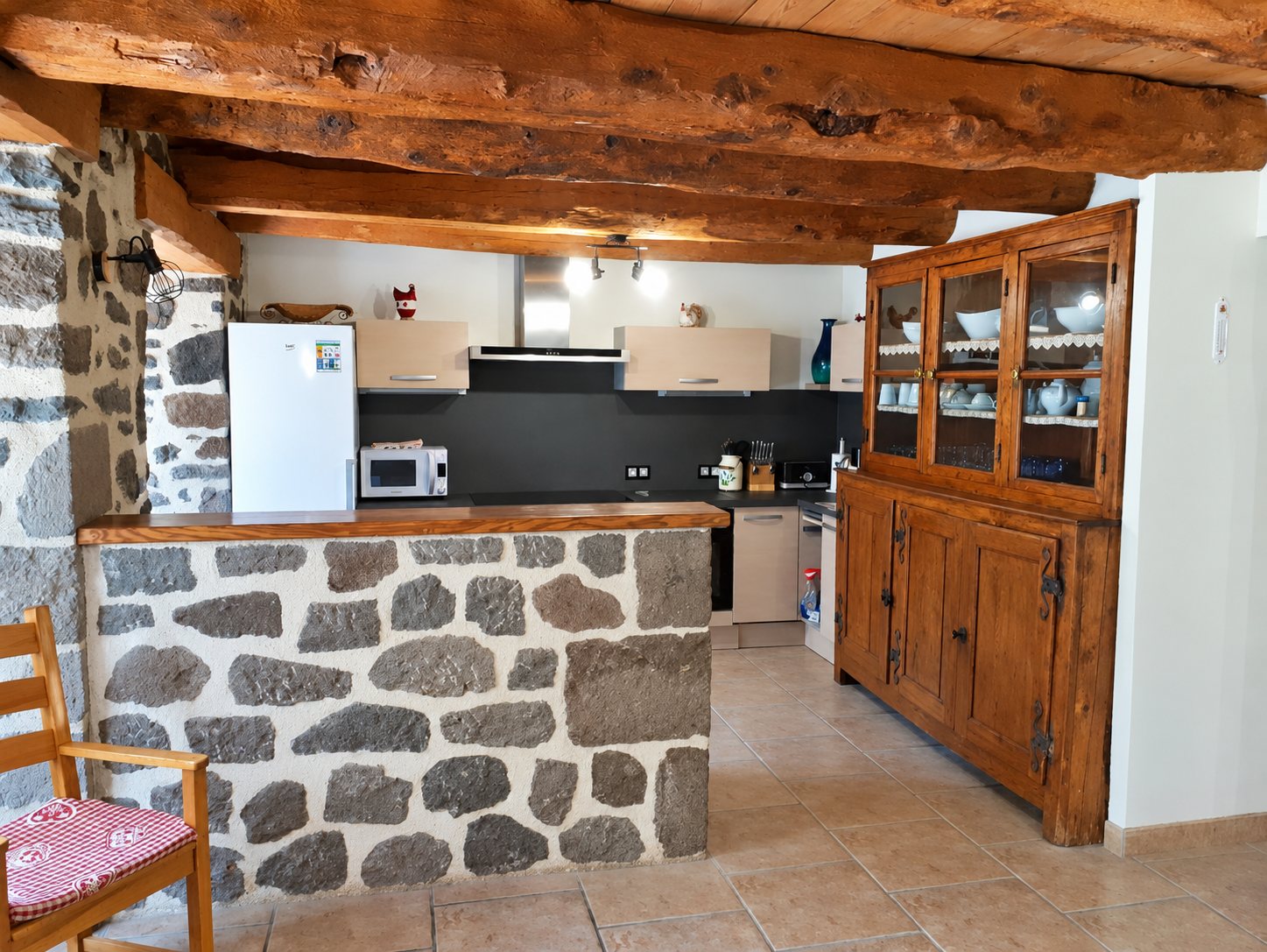 Cuisine ouverte de style rustique avec poutres en bois, comptoir en pierres volcaniques, réfrigérateur blanc et grand vaisselier en bois.