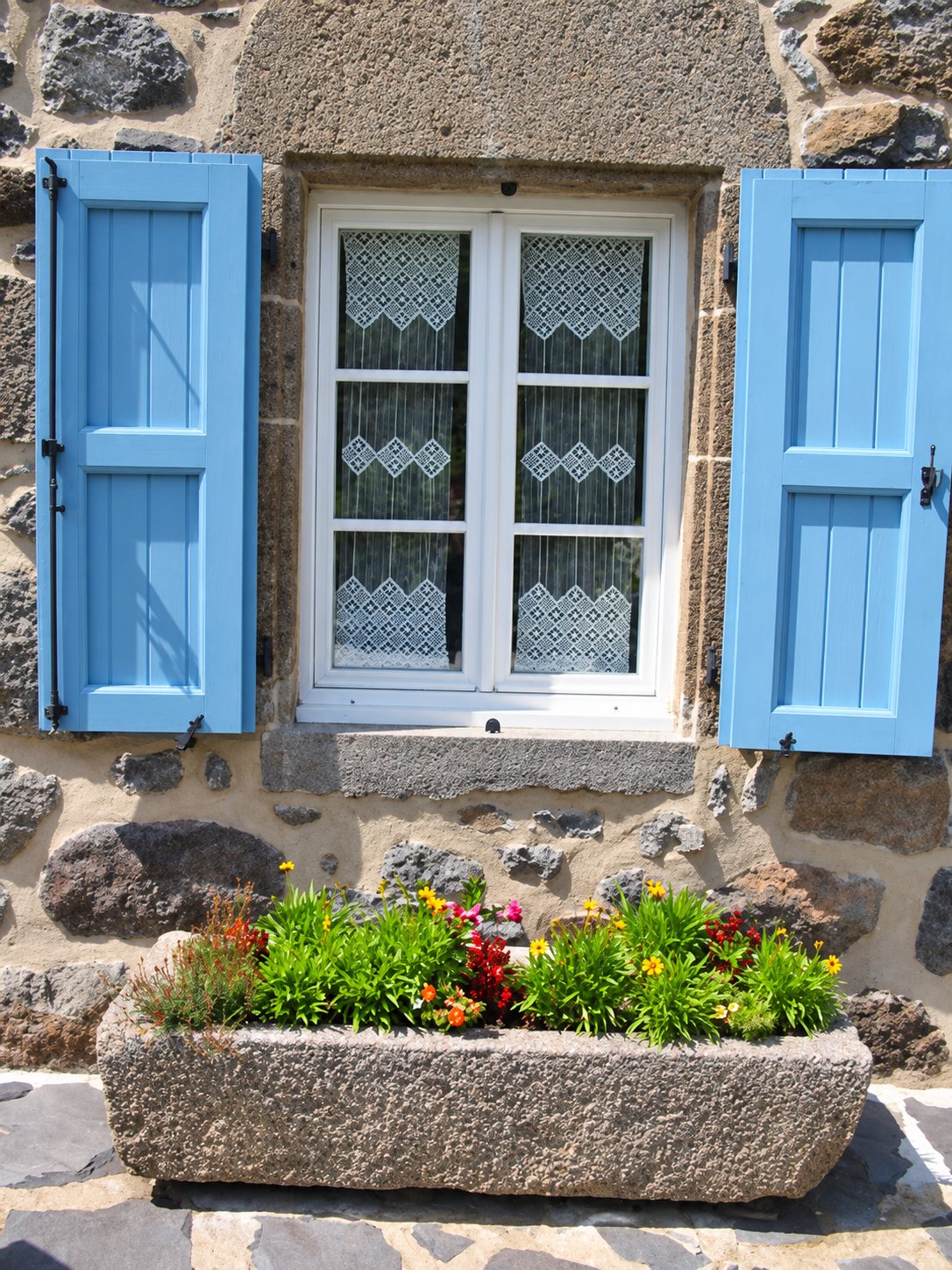 Une fenêtre blanche à rideaux de dentelle encadrée de volets bleu clair sur un mur en pierre, avec une auge en pierre remplie de fleurs au premier plan.