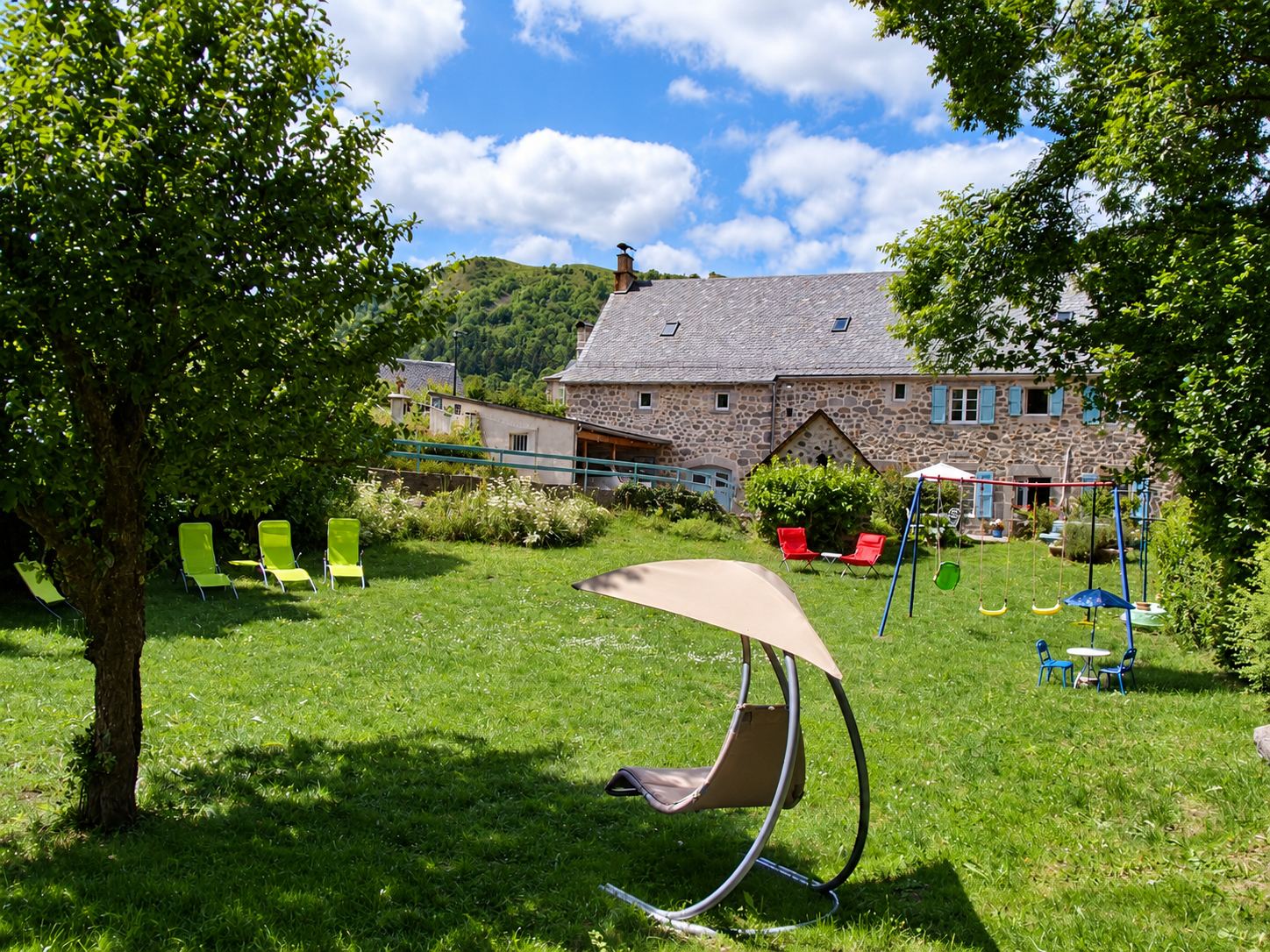 Un jardin herbeux avec des transats verts et rouges, une balançoire pour enfants et un fauteuil suspendu devant une ancienne maison en pierre aux volets bleus, au pied d'une colline boisée.