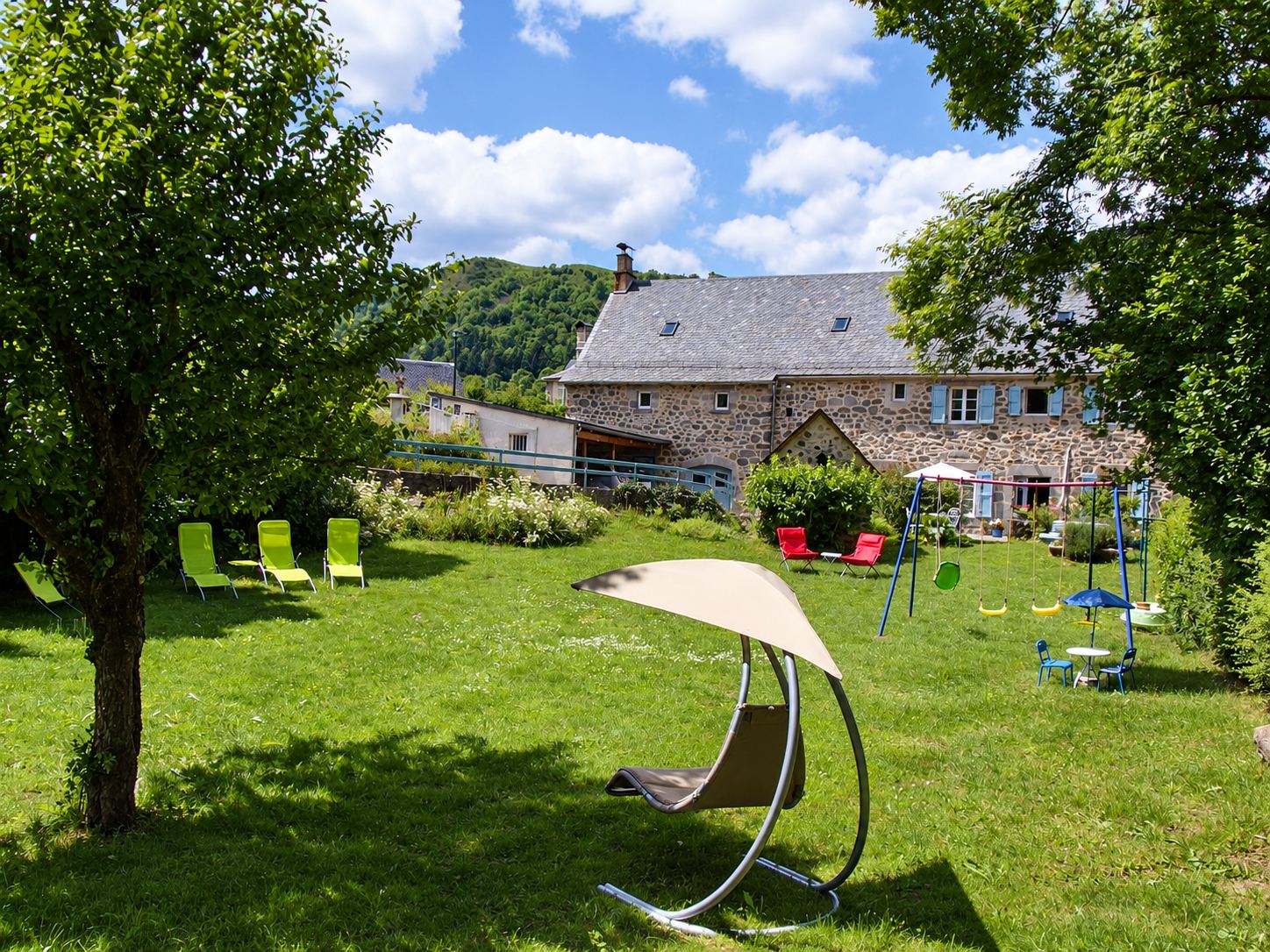 Un jardin herbeux avec transats verts, fauteuils rouges, balançoires et fauteuil suspendu devant une grande maison en pierre aux volets bleus, entourée de collines boisées.