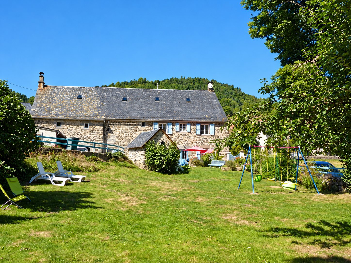 Une ancienne maison en pierre au toit de lauzes, entourée d'un jardin herbeux avec transats et balançoires, au pied d'une colline boisée.