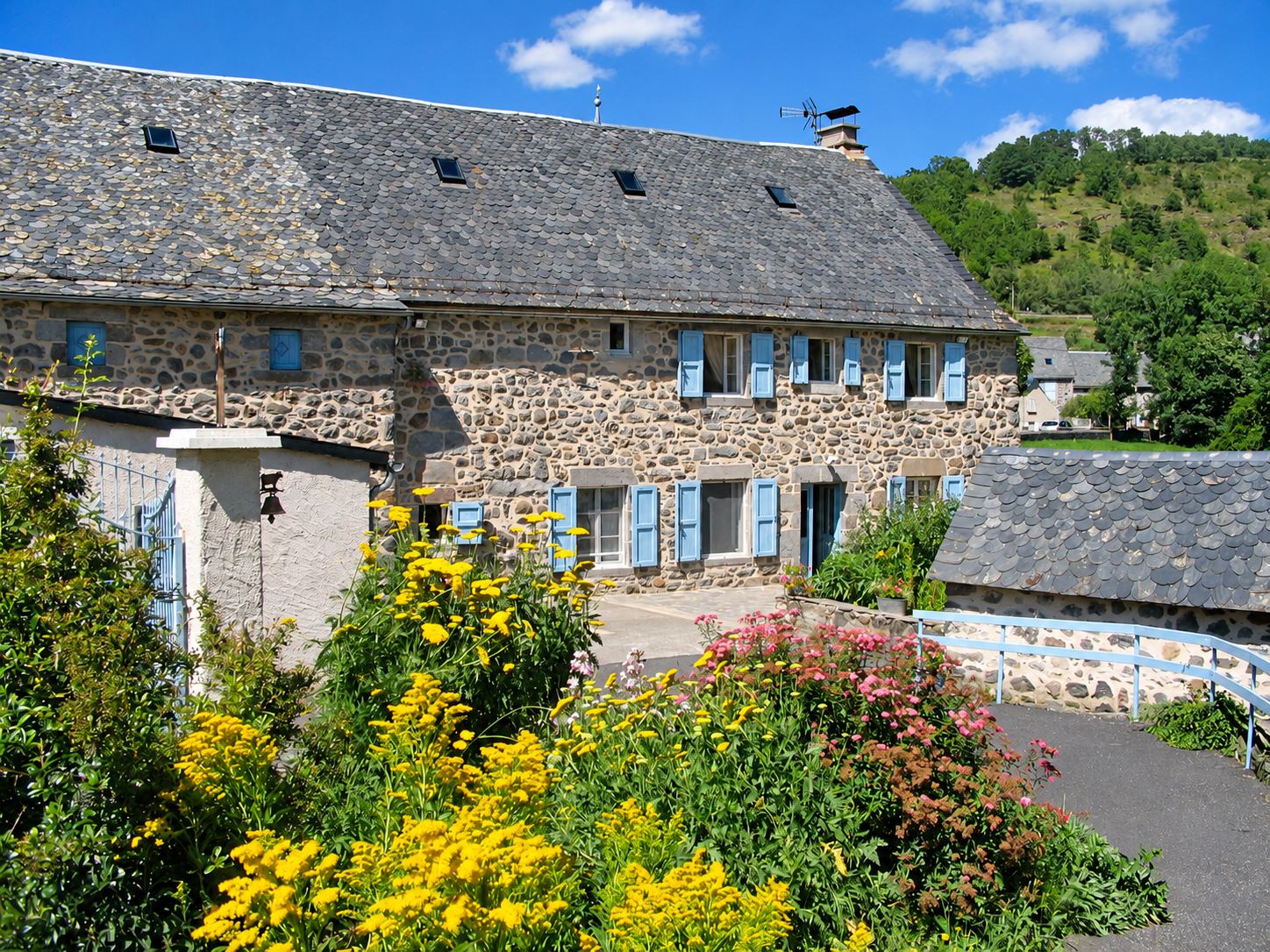 Une ancienne maison en pierre au toit de lauzes et aux volets bleus, devant un jardin fleuri de fleurs jaunes et roses, avec une colline boisée en arrière-plan.