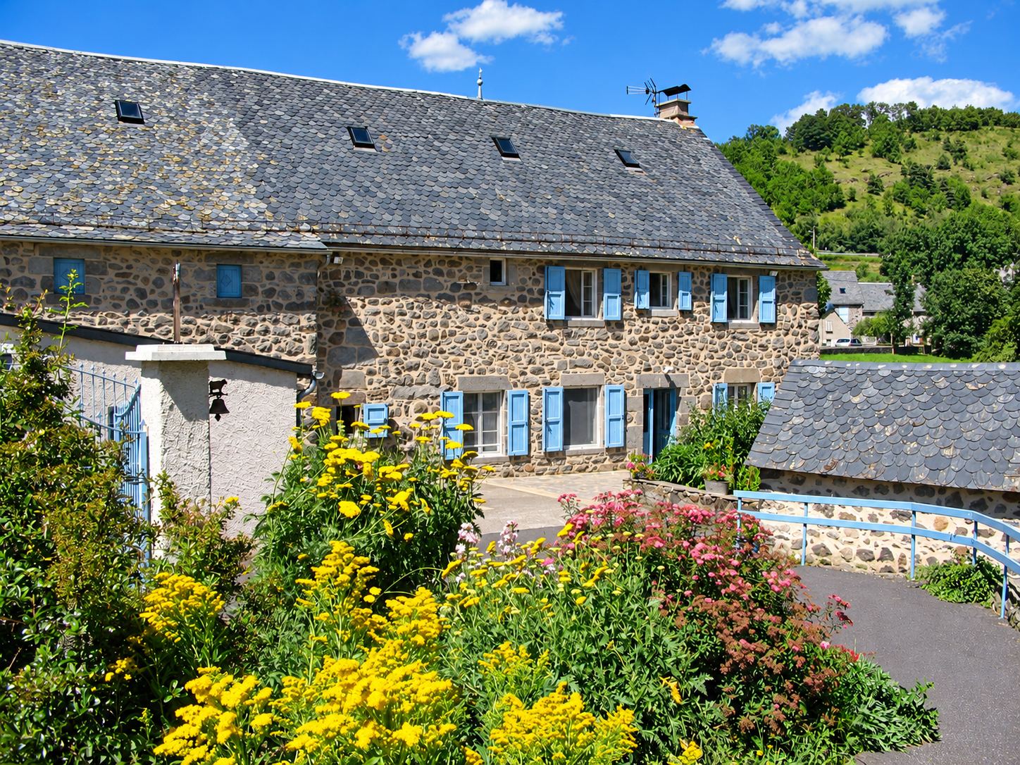 Ancienne maison en pierre au toit de lauzes et aux volets bleus, devant un massif de fleurs jaunes et roses, avec une colline boisée en arrière-plan.