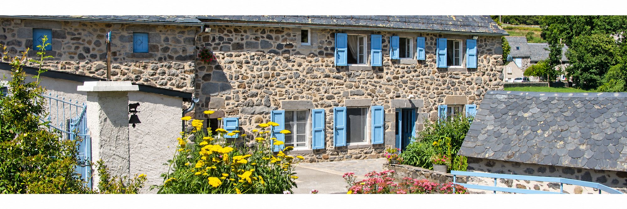 Une maison traditionnelle en pierre avec des volets bleus et un toit de lauzes, entourée de fleurs jaunes et roses dans un cadre verdoyant.