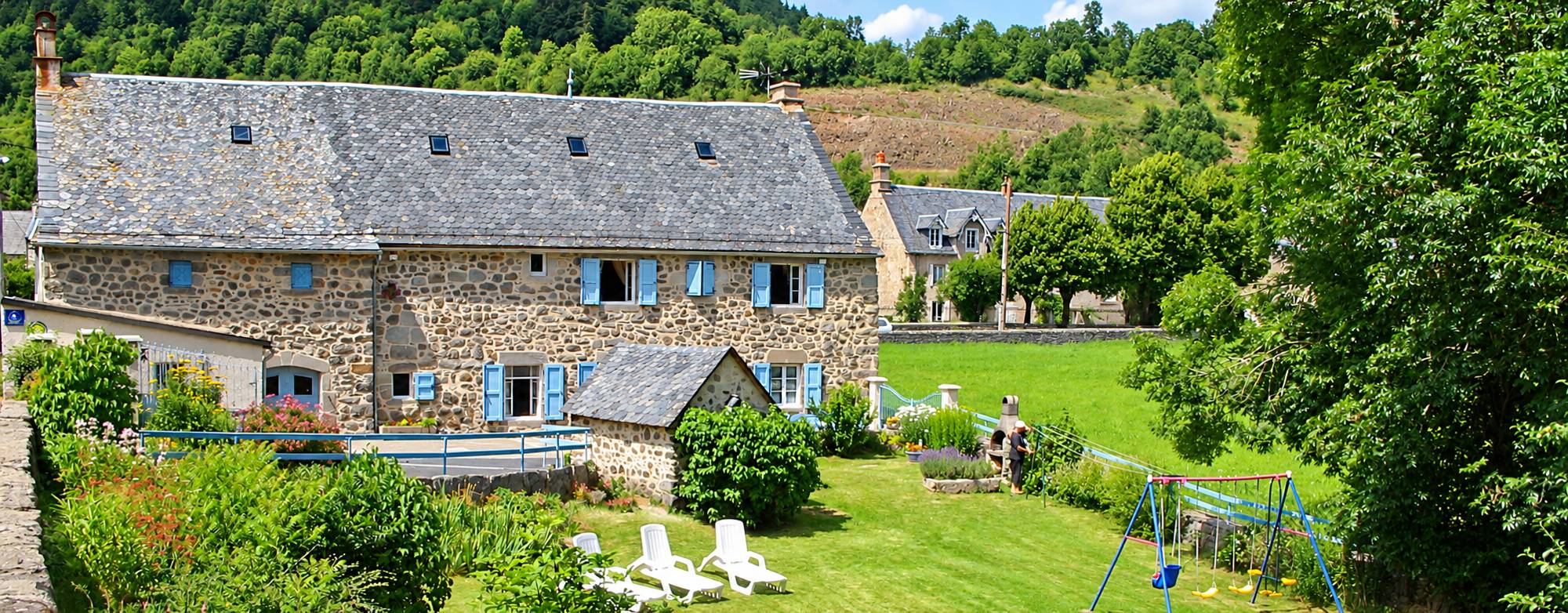 Une ancienne maison en pierre au toit de lauzes et aux volets bleus, entourée d'un jardin avec transats et balançoire, au pied d'une colline boisée.