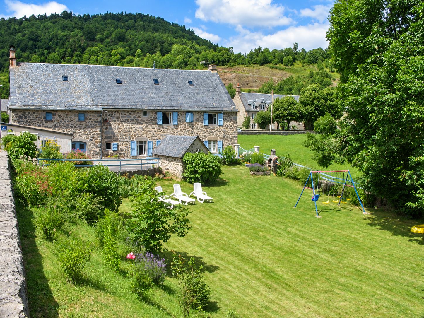 Une grande maison en pierre au toit d'ardoise et aux volets bleus domine un jardin en pente avec transats et balançoire, au pied d'une colline boisée.
