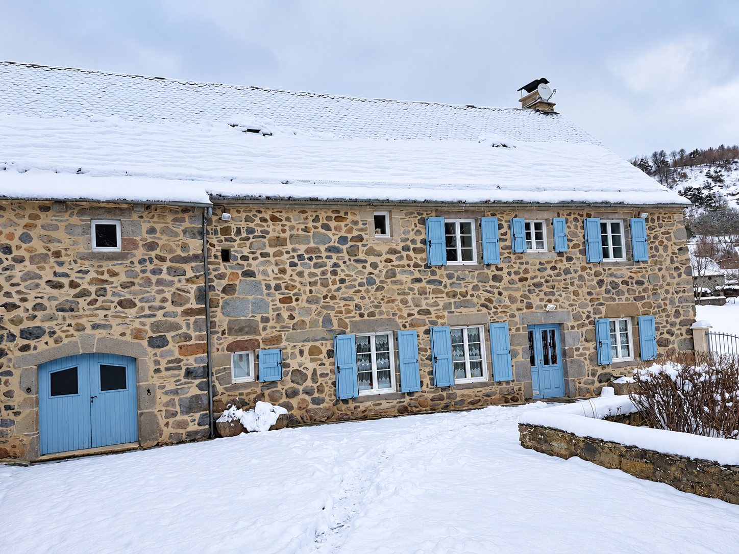 Une longue maison en pierre apparente aux volets et portes bleus, entourée de neige, avec un toit enneigé et une colline boisée à l'arrière-plan.