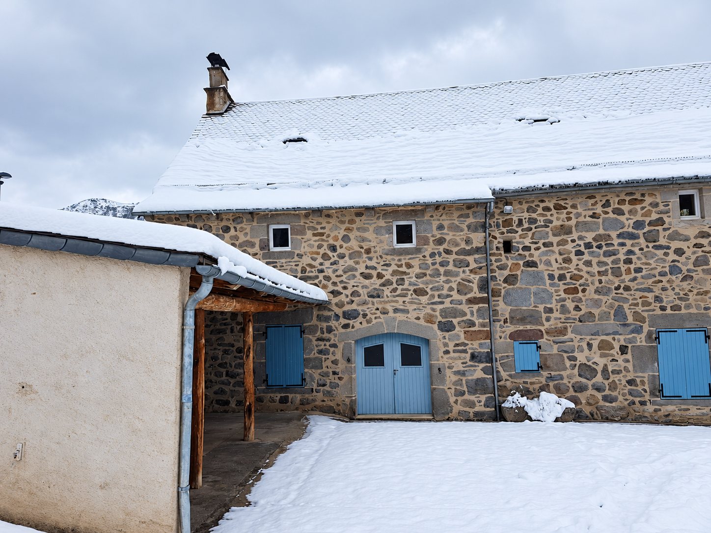 Une maison en pierre au toit enneigé, dotée de volets et d'une porte bleu clair, avec une cour couverte de neige au premier plan.