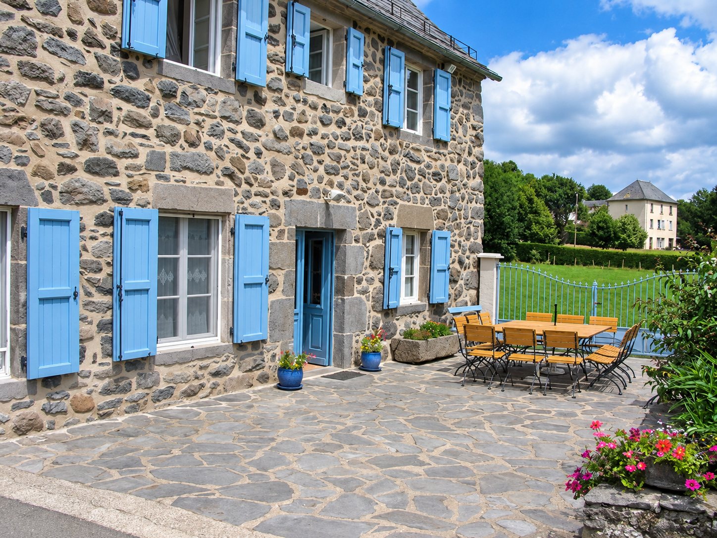 Façade d'une maison en pierre aux volets bleus donnant sur une terrasse dallée avec table, chaises et fleurs, devant un pré verdoyant.