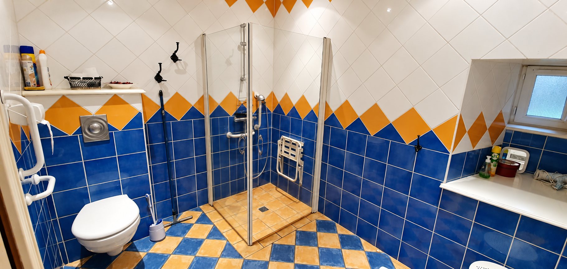 Salle de bain au carrelage bleu et jaune avec une douche à l'italienne équipée d'un siège rabattable, de barres d'appui et d'un WC suspendu.