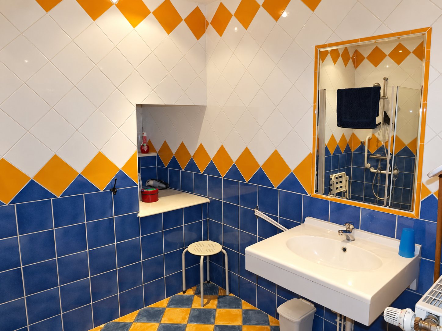 Salle de bain aux murs carrelés de losanges orange et bleus, avec un grand lavabo blanc, un miroir, un tabouret et une douche équipée d'un siège rabattable.