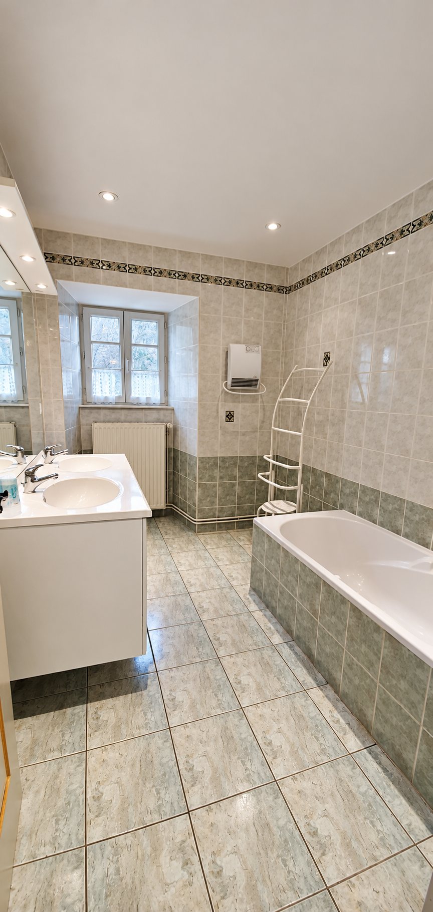 Salle de bain aux murs et au sol carrelés de teintes grises et vertes, avec meuble à double vasque, baignoire, radiateur et fenêtre.