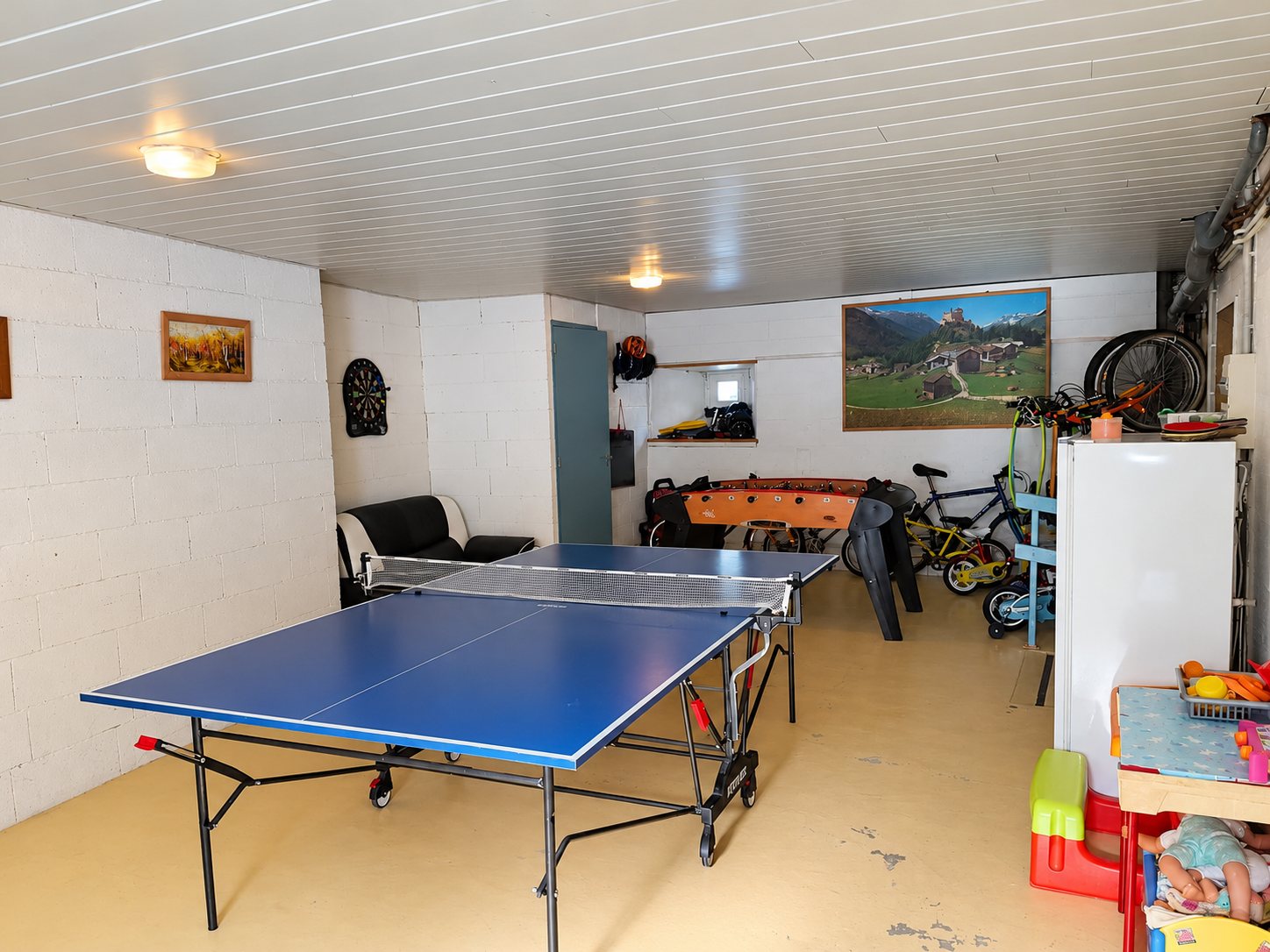 Une salle de jeux aux murs blancs avec une table de ping-pong bleue, un baby-foot, un canapé noir, une cible de fléchettes et des vélos d'enfants.