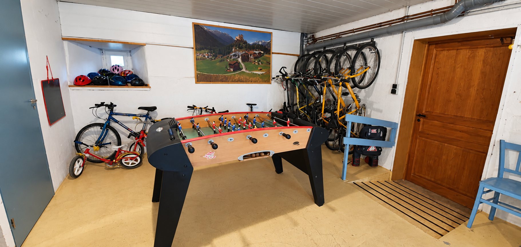 Une salle au sol jaune avec un baby-foot au centre, des vélos accrochés au mur de droite et des casques posés sur une étagère.