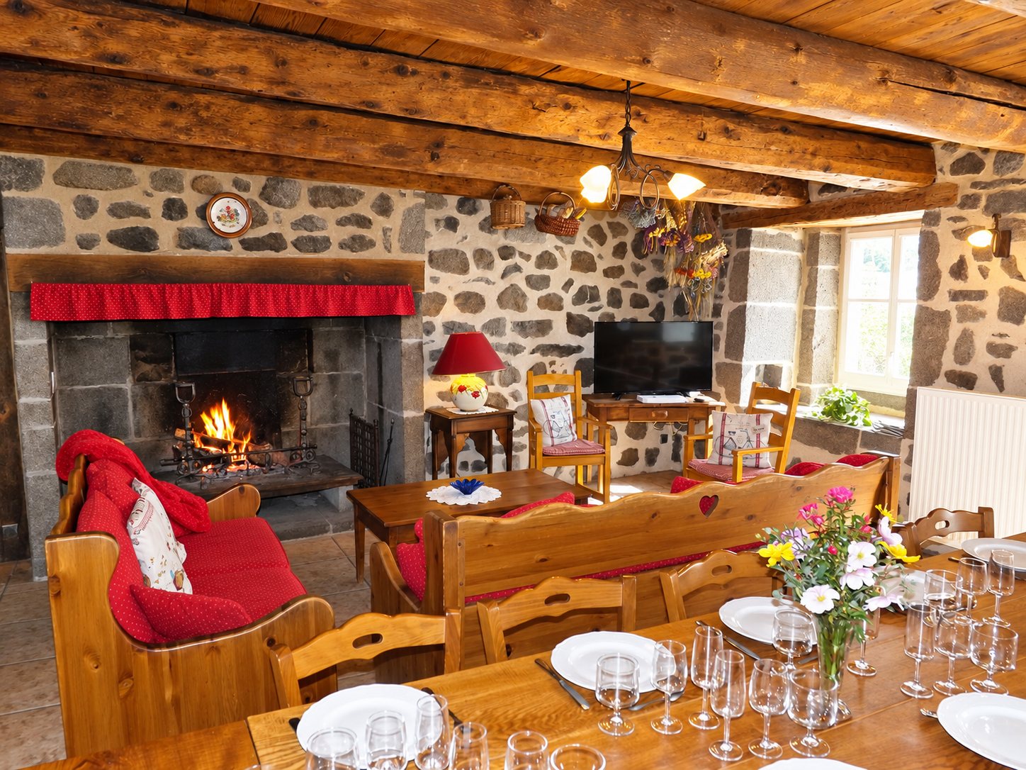 Salle à manger rustique aux murs de pierre et aux poutres apparentes, avec une cheminée allumée et une grande table dressée avec assiettes et verres.