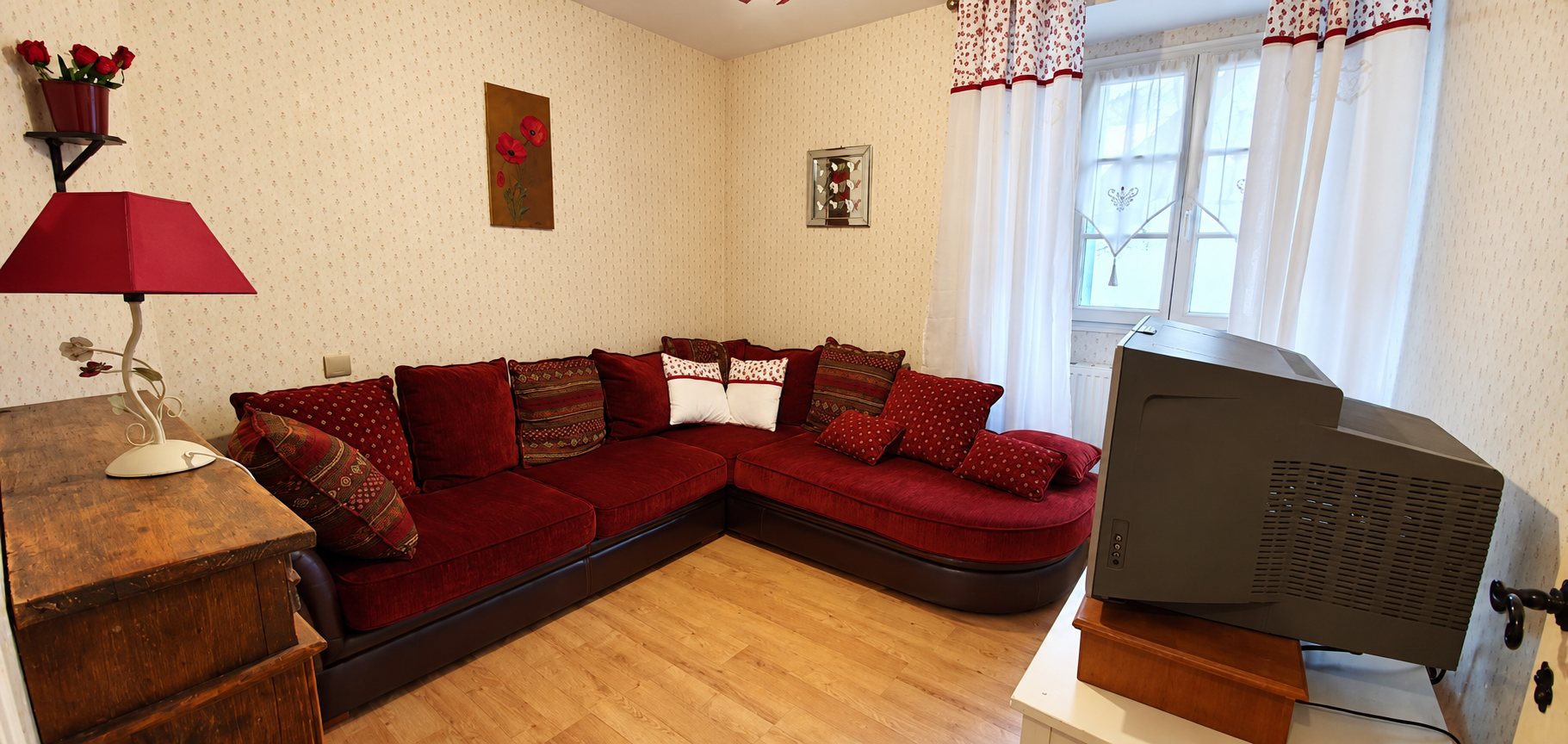 Salon aux murs tapissés de beige avec un grand canapé d'angle rouge garni de coussins, une lampe rouge, une fenêtre aux rideaux blancs et un ancien téléviseur.