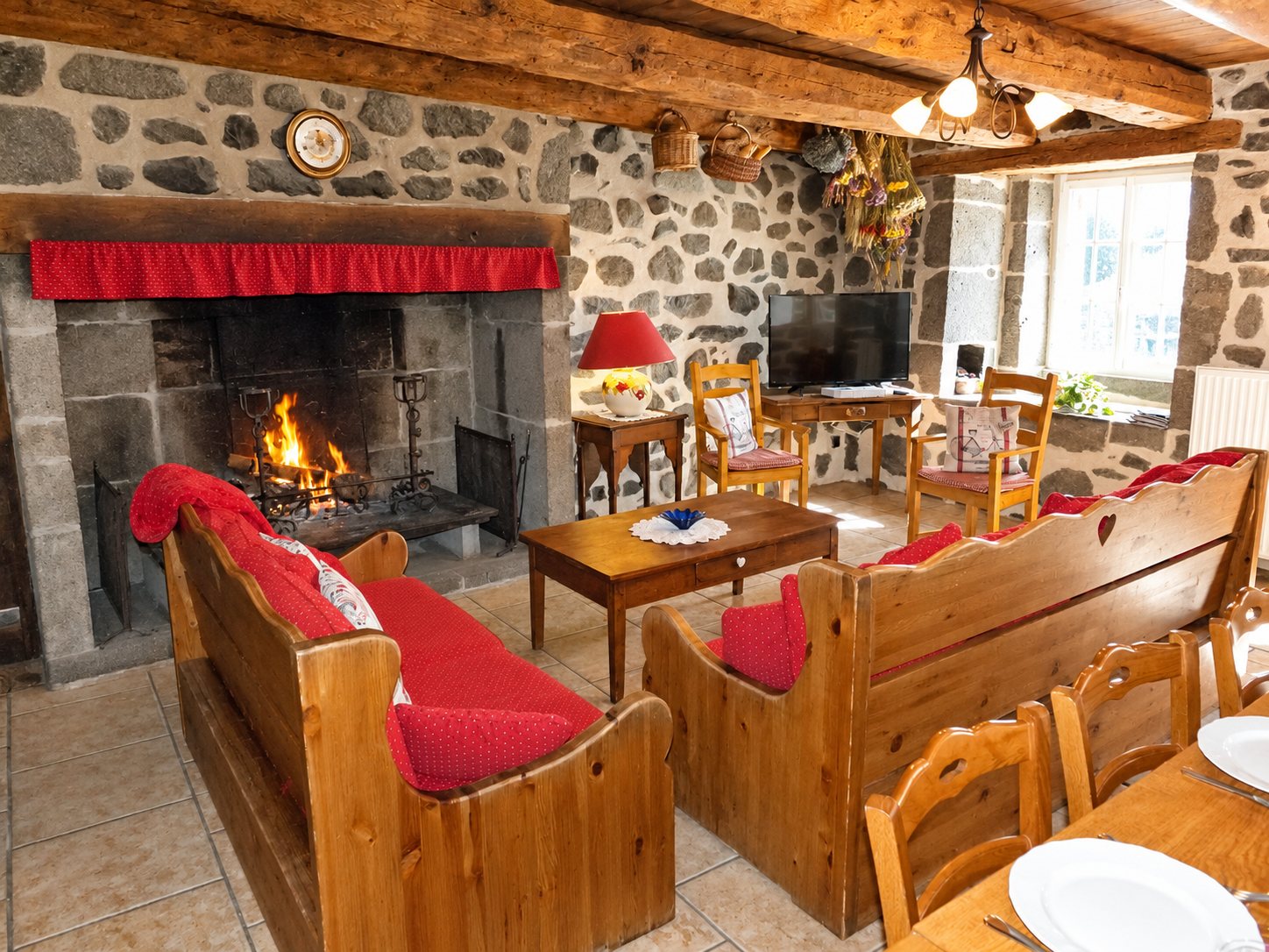 Salon d'un gîte aux murs de pierre avec une cheminée où brûle un feu, des banquettes en bois à coussins rouges, une table basse et une table dressée au premier plan.