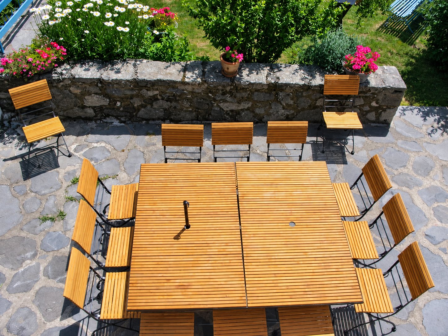 Vue de dessus d'une grande table carrée en lattes de bois entourée de chaises pliantes en bois et métal sur une terrasse dallée de pierre, avec des fleurs et un muret en arrière-plan.