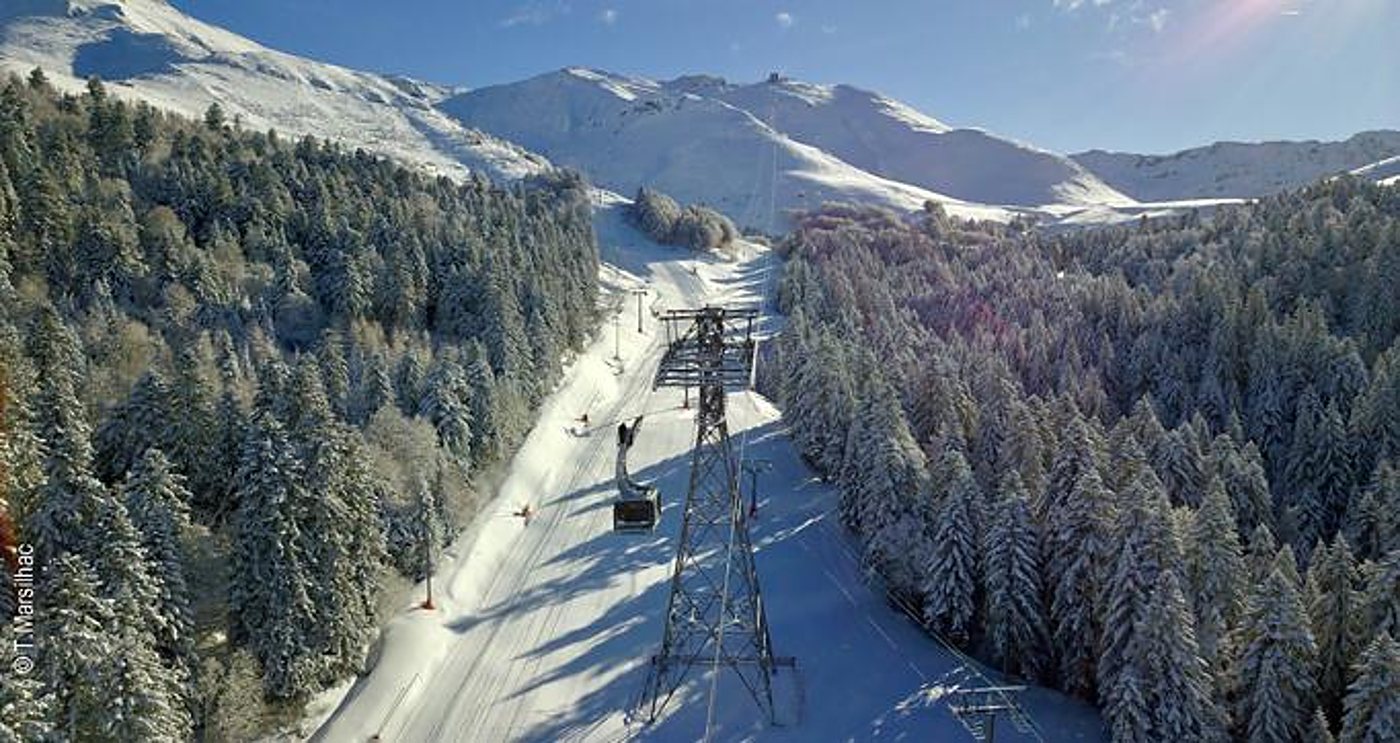 Vue aérienne d'une télécabine suspendue à un pylône métallique au-dessus d'une piste de ski enneigée bordée de sapins givrés, avec des montagnes blanches au fond.