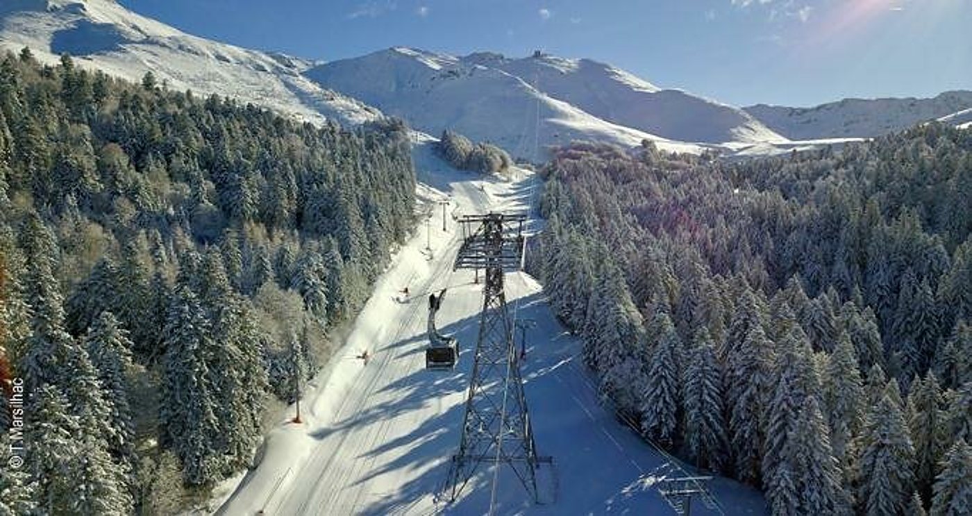 Vue aérienne d'une télécabine et de son pylône au-dessus d'une piste de ski enneigée bordée de sapins couverts de neige, avec des montagnes en arrière-plan.