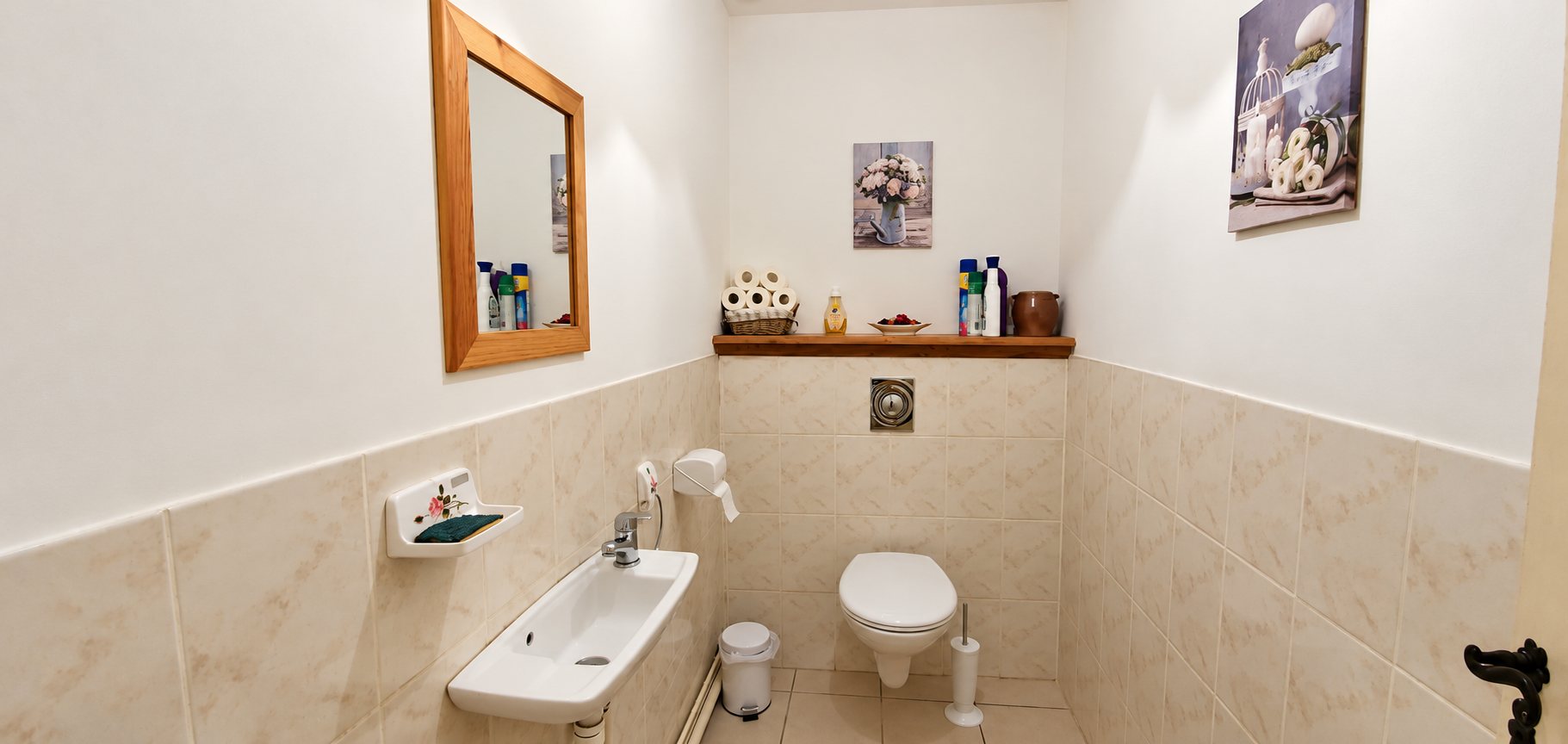 Des toilettes aux murs blancs et carrelage beige avec un petit lavabo, un miroir encadré de bois, une étagère décorée et des tableaux floraux.